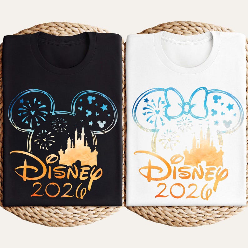 May include: A black t-shirt and a white t-shirt, each featuring a graphic of a castle silhouette inside a Mickey Mouse ear outline with fireworks, stars, and the text Disney 2026. The white shirt includes a bow on the Mickey ear outline.