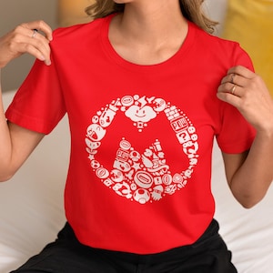 May include: A red crew neck t-shirt featuring a white circular design with various cartoon characters and icons. The design includes a cloud, question mark block, and other elements from a popular video game.
