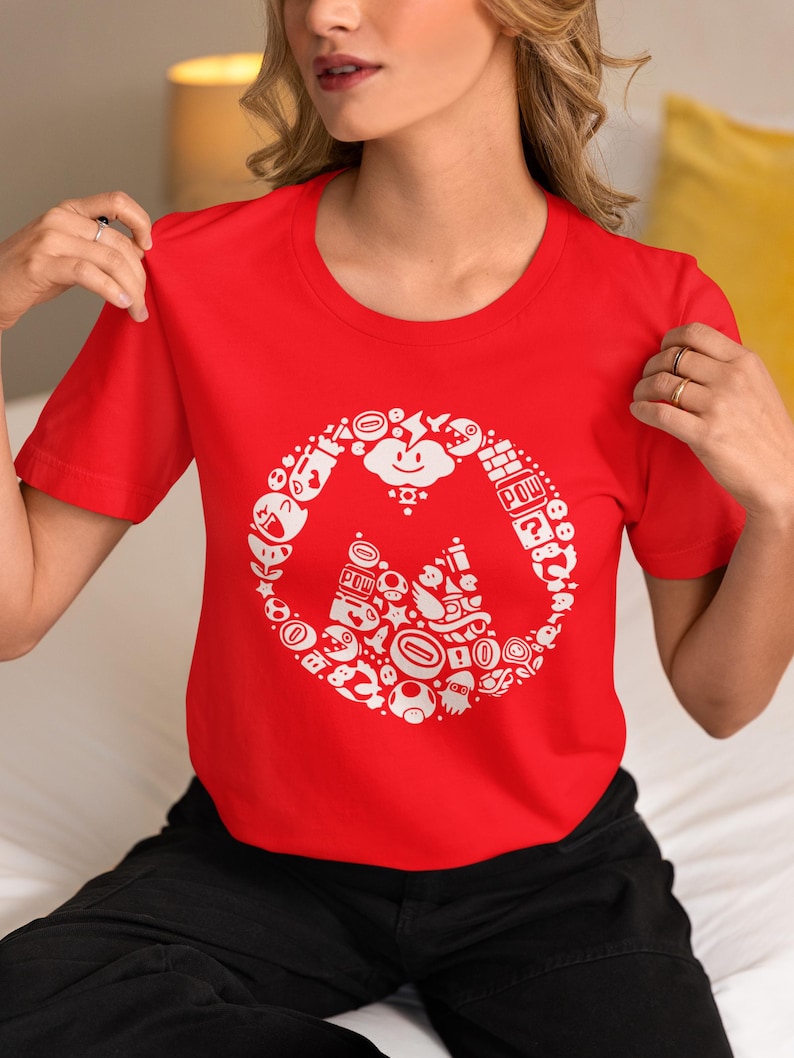 May include: A red crew neck t-shirt featuring a white circular design with various cartoon characters and icons. The design includes a cloud, question mark block, and other elements from a popular video game.