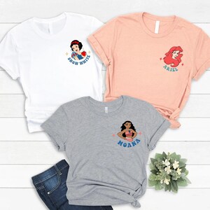 Op de afbeelding: Drie t-shirts in wit, perzik en grijs, elk met een cartoonkarakterontwerp. Het witte shirt heeft Sneeuwwitje, het perzikkleurige shirt heeft Ariel en het grijze shirt heeft Moana. Elk ontwerp bevat de naam van het personage.
