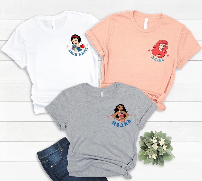 Op de afbeelding: Drie t-shirts in wit, perzik en grijs, elk met een cartoonkarakterontwerp. Het witte shirt heeft Sneeuwwitje, het perzikkleurige shirt heeft Ariel en het grijze shirt heeft Moana. Elk ontwerp bevat de naam van het personage.