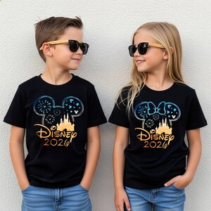 May include: Two black t-shirts featuring a graphic of a castle silhouette inside a Mickey Mouse ear outline with fireworks and stars. The text reads Disney 2026 in a gradient orange font. One shirt includes a Minnie Mouse bow on the ear outline.