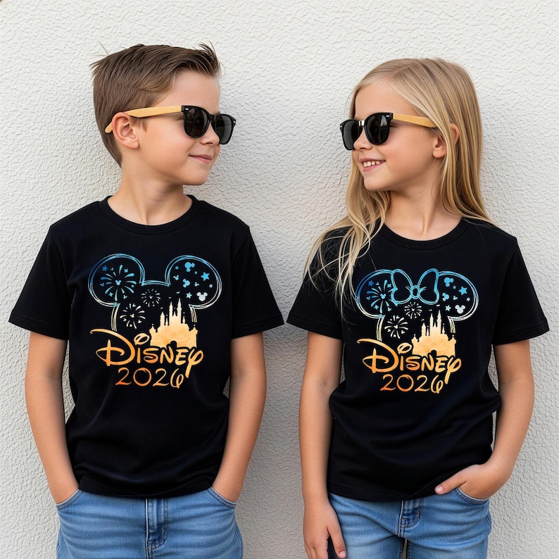 May include: Two black t-shirts featuring a graphic of a castle silhouette inside a Mickey Mouse ear outline with fireworks and stars. The text reads Disney 2026 in a gradient orange font. One shirt includes a Minnie Mouse bow on the ear outline.