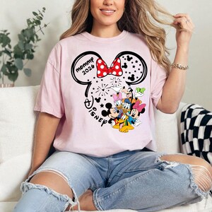 Puede incluir: Camiseta rosa claro con un dise&ntilde;o de Minnie Mouse, un lazo rojo de lunares y el texto "Mommy 2026". El dise&ntilde;o incluye personajes de Disney y la palabra "Disney".