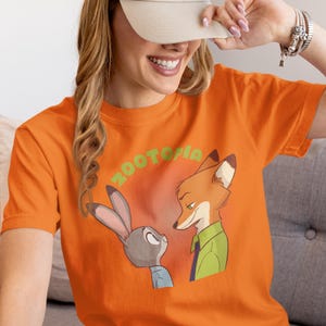 May include: Orange t-shirt featuring a graphic of Judy Hopps and Nick Wilde from the film Zootopia. The text "Zootopia" is printed in green letters above the characters.