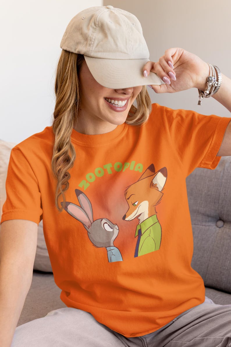 May include: Orange t-shirt featuring a graphic of Judy Hopps and Nick Wilde from the film Zootopia. The text "Zootopia" is printed in green letters above the characters.