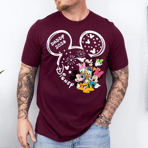 Puede incluir: Camiseta burdeos con un dise&ntilde;o blanco de la cabeza de Mickey Mouse, el texto "DADDY 2026" y la palabra "Disney". El dise&ntilde;o incluye fuegos artificiales y estrellas, con los personajes de Mickey, Minnie, Donald, Daisy, Goofy y Pluto.