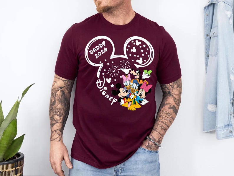 Puede incluir: Camiseta burdeos con un dise&ntilde;o blanco de la cabeza de Mickey Mouse, el texto "DADDY 2026" y la palabra "Disney". El dise&ntilde;o incluye fuegos artificiales y estrellas, con los personajes de Mickey, Minnie, Donald, Daisy, Goofy y Pluto.