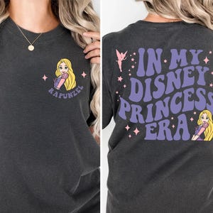 Op de afbeelding: Donkergrijs t-shirt met een cartoon illustratie van Rapunzel en de tekst "Rapunzel" op de voorkant. De achterkant van het shirt leest "In My Disney Princess Era" in paars, met een fee en een Rapunzel graphic.