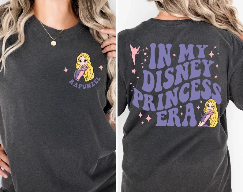 Op de afbeelding: Donkergrijs t-shirt met een cartoon illustratie van Rapunzel en de tekst "Rapunzel" op de voorkant. De achterkant van het shirt leest "In My Disney Princess Era" in paars, met een fee en een Rapunzel graphic.
