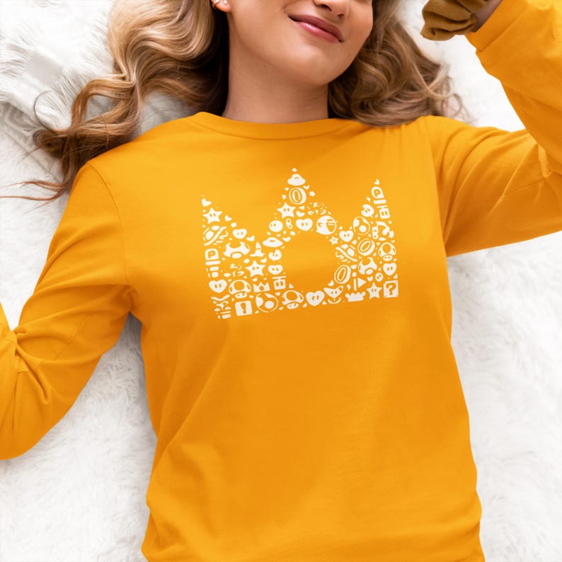 May include: Mustard yellow long-sleeve shirt featuring a white crown design filled with various video game icons. The crown is centered on the chest. The shirt is made of a soft material.
