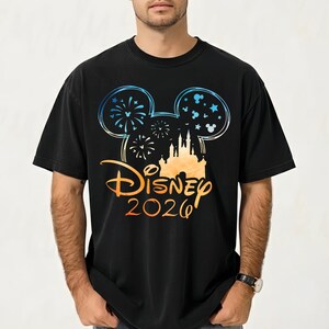 May include: A black t-shirt featuring a graphic of a Mickey Mouse ear silhouette filled with fireworks and stars. Below the ears is a castle illustration, the word Disney in a script font, and the year 2026 in a gradient orange and blue color.