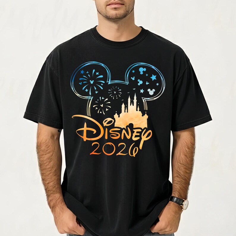 May include: A black t-shirt featuring a graphic of a Mickey Mouse ear silhouette filled with fireworks and stars. Below the ears is a castle illustration, the word Disney in a script font, and the year 2026 in a gradient orange and blue color.