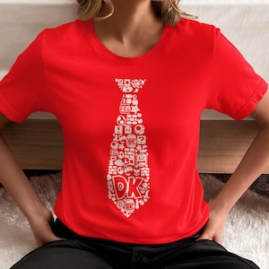 May include: A red t-shirt featuring a white tie design composed of various video game icons and the letters "DK". The short-sleeved shirt is made of a soft material and is suitable for casual wear. The design is a playful nod to classic video games.
