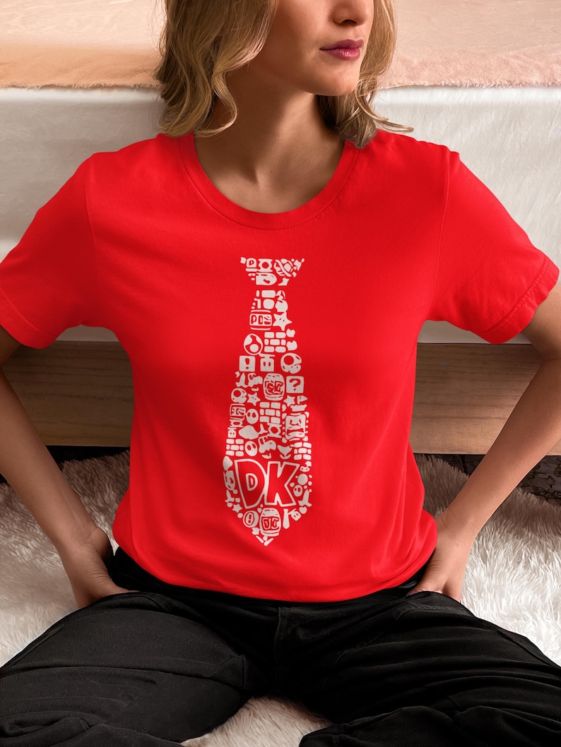 May include: A red t-shirt featuring a white tie design composed of various video game icons and the letters "DK". The short-sleeved shirt is made of a soft material and is suitable for casual wear. The design is a playful nod to classic video games.