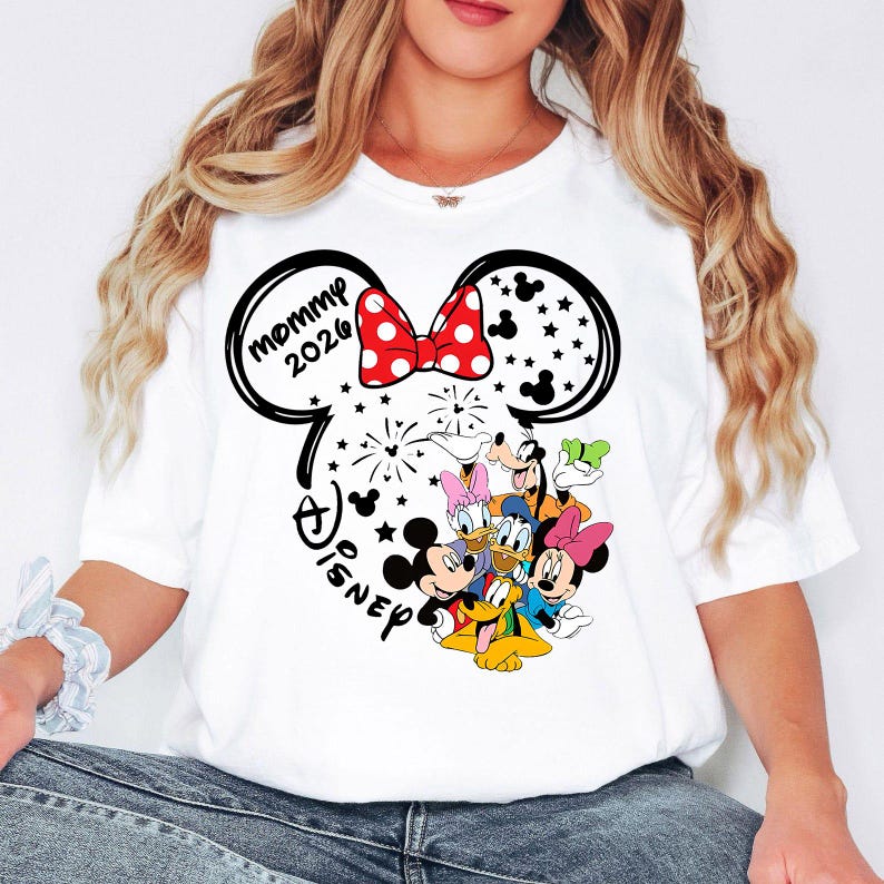 Puede incluir: Camiseta blanca con un dise&ntilde;o de orejas de Mickey Mouse, un lazo rojo con lunares y el texto "mommy 2026". El dise&ntilde;o incluye personajes de Disney como Mickey, Minnie, Donald y Goofy.