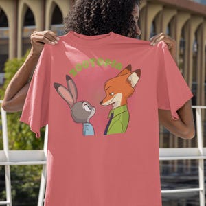 May include: A pink t-shirt featuring a cartoon image of a fox and a rabbit looking at each other. The text "Zootopia" is printed in green on the shirt.