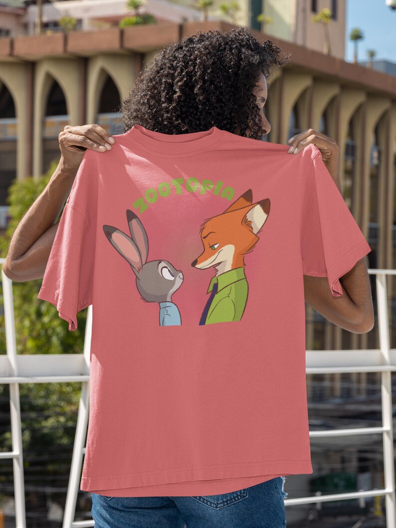 May include: A pink t-shirt featuring a cartoon image of a fox and a rabbit looking at each other. The text "Zootopia" is printed in green on the shirt.