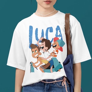 May include: White t-shirt with a blue graphic of three cartoon characters riding a scooter. The characters are smiling and the text "LUCA" is printed in blue above them.