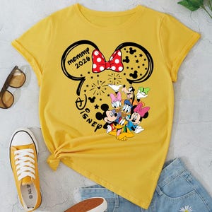 Puede incluir: Camiseta amarilla con dise&ntilde;o de Minnie Mouse, texto "mommy 2026" y personajes de Disney. La imagen incluye pantalones cortos de mezclilla y zapatillas amarillas. Tambi&eacute;n se ven gafas de sol.