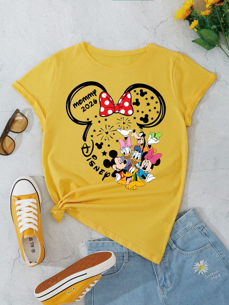 Puede incluir: Camiseta amarilla con dise&ntilde;o de Minnie Mouse, texto "mommy 2026" y personajes de Disney. La imagen incluye pantalones cortos de mezclilla y zapatillas amarillas. Tambi&eacute;n se ven gafas de sol.