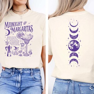 May include: A cream-colored t-shirt featuring a purple graphic on the front with the text Midnight Margaritas, a margarita glass, a frog wearing a hat, a coffee pot, and small botanical elements. The back displays a vertical purple moon phase design.