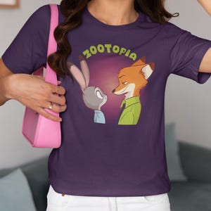 May include: Purple t-shirt featuring a graphic of Judy Hopps and Nick Wilde from the film Zootopia. The characters are looking at each other with the text "Zootopia" above them.