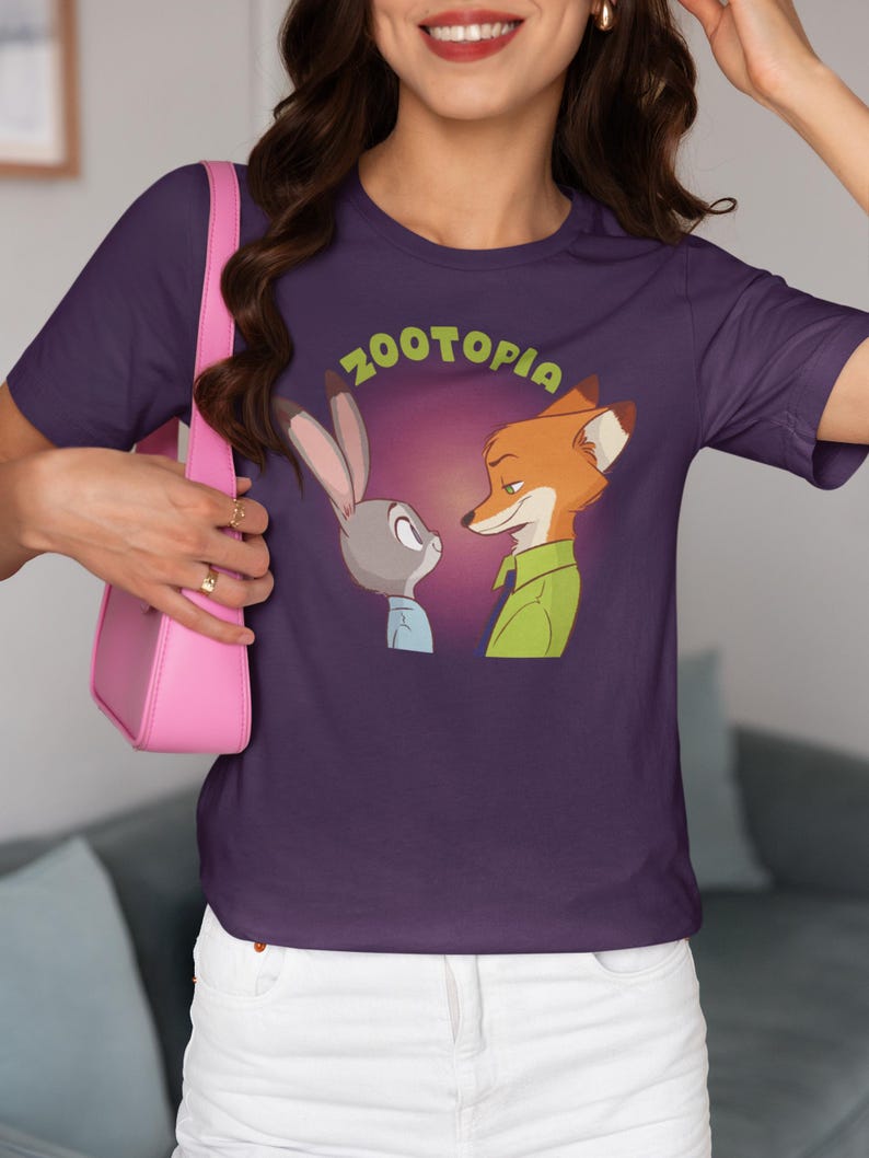 May include: Purple t-shirt featuring a graphic of Judy Hopps and Nick Wilde from the film Zootopia. The characters are looking at each other with the text "Zootopia" above them.