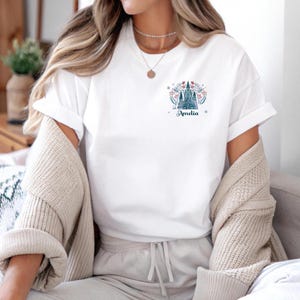 Op de afbeelding: Wit T-shirt met een kleine, kleurrijke afbeelding van een kasteel, bloemen en de naam "Amelia" in een sierlijk lettertype. Het shirt heeft korte mouwen en een relaxte pasvorm. Een beige vest is over de schouders gedrapeerd.