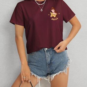 Op de afbeelding: Bordeauxrood T-shirt met een cartoon illustratie van Belle uit Belle en het Beest, met het woord "BELLE" in geel. Het shirt wordt gedragen met een spijkerbroekshort.