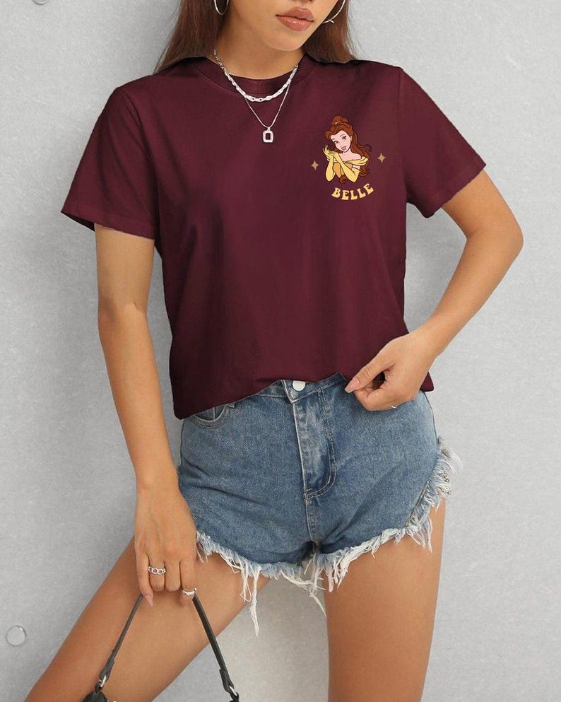Op de afbeelding: Bordeauxrood T-shirt met een cartoon illustratie van Belle uit Belle en het Beest, met het woord "BELLE" in geel. Het shirt wordt gedragen met een spijkerbroekshort.