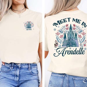 Op de afbeelding: Cr&egrave;mekleurig T-shirt met een ontwerp op de voor- en achterkant. De voorkant heeft een kasteelafbeelding en de naam "Amelia". De achterkant toont de tekst "Meet Me In Arendelle" met een kasteelillustratie en bloemmotieven.