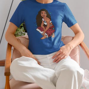 May include: A royal blue t-shirt with a cartoon image of a woman with long brown hair wearing a red and white outfit. The woman is sitting with her legs crossed and has a necklace with a blue gem. The t-shirt has a few small white stars printed on it.