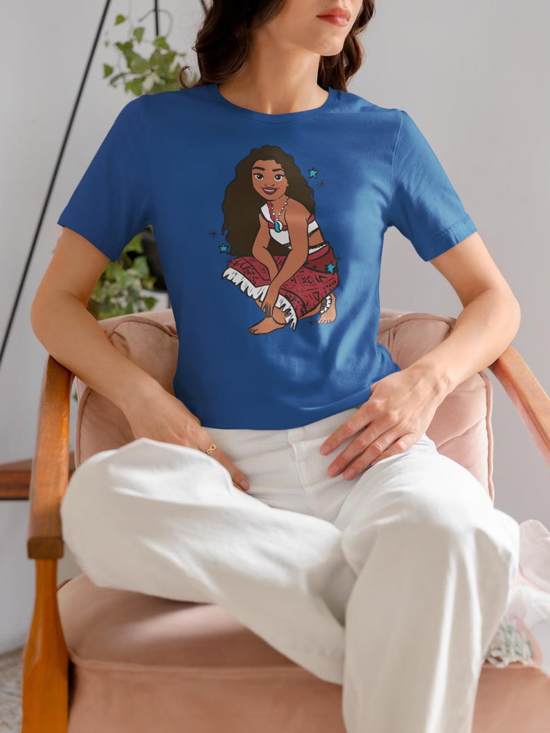 May include: A royal blue t-shirt with a cartoon image of a woman with long brown hair wearing a red and white outfit. The woman is sitting with her legs crossed and has a necklace with a blue gem. The t-shirt has a few small white stars printed on it.