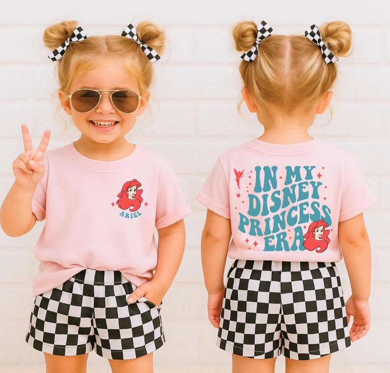 Op de afbeelding: Een jong meisje draagt een roze t-shirt met een Ariel-afbeelding en de tekst "ARIEL". Ze draagt ook een zwart-wit geruite short. De achterkant van het shirt zegt "IN MY DISNEY PRINCESS ERA".
