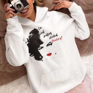 May include: White hooded sweatshirt with a black and white illustration of a woman's face with red lipstick. The text "I'm just getting started, Darling!" is printed on the sweatshirt.
