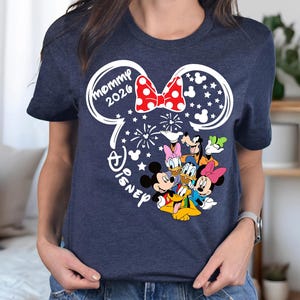 Puede incluir: Camiseta azul marino con un dise&ntilde;o de orejas de Mickey Mouse blancas y un lazo rojo de lunares. El dise&ntilde;o incluye el texto "mommy 2026" y los personajes de Disney Mickey, Minnie, Donald, Daisy, Goofy y Pluto.