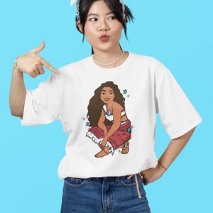 May include: White t-shirt with a cartoon image of a woman with long brown hair wearing a red and white patterned skirt and a blue necklace. The woman is sitting with her legs crossed and has a blue background with white stars.