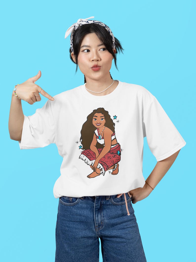 May include: White t-shirt with a cartoon image of a woman with long brown hair wearing a red and white patterned skirt and a blue necklace. The woman is sitting with her legs crossed and has a blue background with white stars.