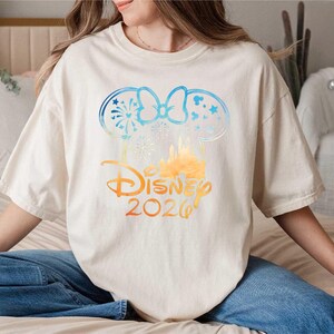 May include: An off-white short-sleeve t-shirt featuring a graphic of mouse ears with a bow, fireworks, and a castle silhouette. The design transitions from blue to orange and includes the text Disney 2026 in a stylized orange font.