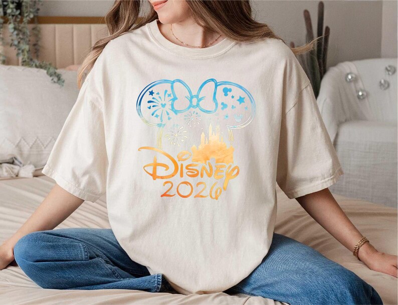 May include: An off-white short-sleeve t-shirt featuring a graphic of mouse ears with a bow, fireworks, and a castle silhouette. The design transitions from blue to orange and includes the text Disney 2026 in a stylized orange font.