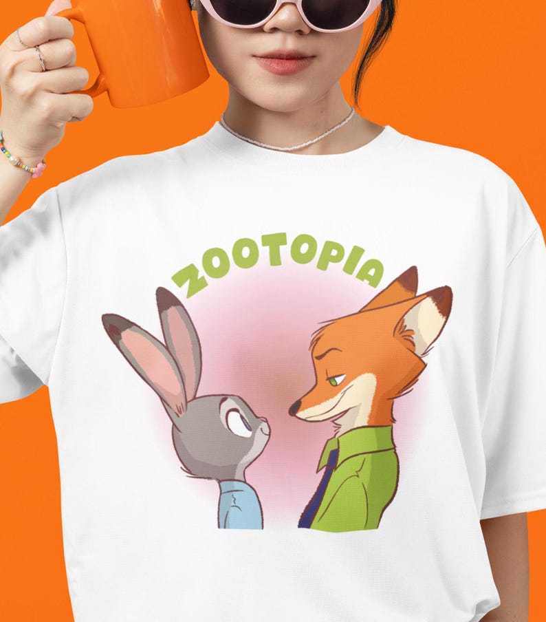 May include: White t-shirt featuring a graphic of Judy Hopps and Nick Wilde from the film Zootopia. The text "Zootopia" is written in green above the characters.