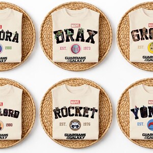 May include: Cream-colored t-shirts featuring Marvel's Guardians of the Galaxy characters: Gamora, Drax, Groot, Star-Lord, Rocket, and Yondu. Each shirt displays the character's name, a logo, and the year of establishment. The shirts are presented on woven circular mats.