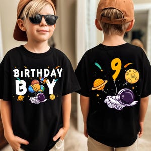 May include: Black t-shirt with a colorful space-themed birthday design. The front features the words "BIRTHDAY BOY" with planets, a rocket, and an astronaut. The back displays a number 9 balloon and an astronaut.