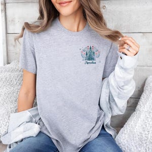 Op de afbeelding: Lichtgrijs t-shirt met een kasteelontwerp, bloemendetails en de naam "Amelia" in teal. Het ontwerp staat op de linkerborst. Het shirt heeft een ronde hals.