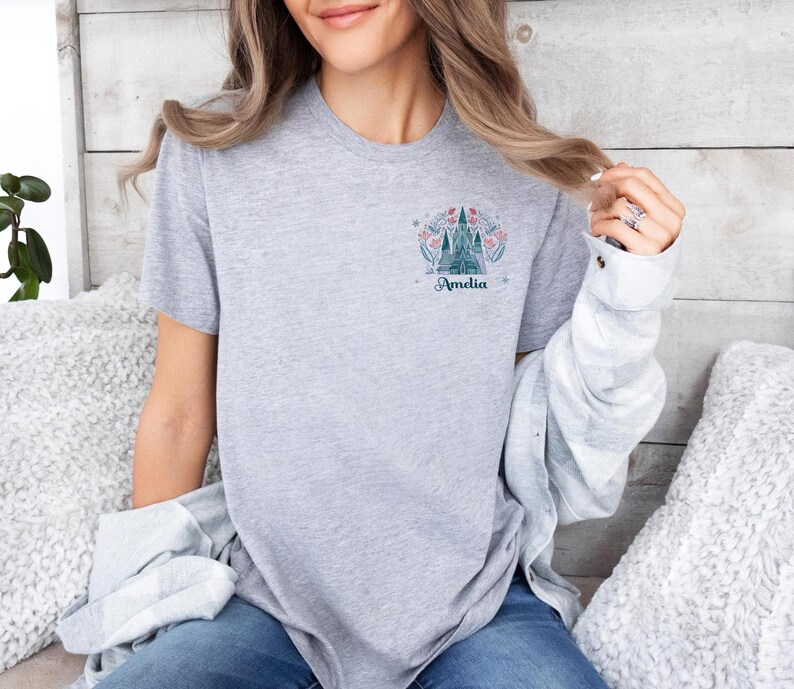 Op de afbeelding: Lichtgrijs t-shirt met een kasteelontwerp, bloemendetails en de naam "Amelia" in teal. Het ontwerp staat op de linkerborst. Het shirt heeft een ronde hals.