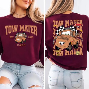 Pode incluir: Camisola bord&ocirc; com uma imagem de desenho animado do Mate do filme Carros. O design frontal inclui o texto "TOW MATER" acima do personagem, com "EST. 2006" e "CARS" abaixo. O design traseiro repete o personagem e o texto.