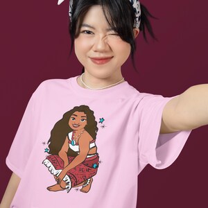 May include: Pink t-shirt with a cartoon illustration of a brown-haired woman wearing a red and white patterned skirt and a blue necklace. The woman is sitting with her legs crossed and has a blue star above her head.
