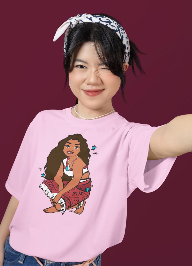 May include: Pink t-shirt with a cartoon illustration of a brown-haired woman wearing a red and white patterned skirt and a blue necklace. The woman is sitting with her legs crossed and has a blue star above her head.