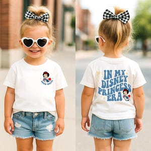 Op de afbeelding: Cr&egrave;mekleurig T-shirt met een Sneeuwwitje-afbeelding op de voorkant en de tekst "IN MY DISNEY PRINCESS ERA" op de achterkant. Het kind draagt een spijkerbroekshort en hartvormige zonnebril, met een zwart-wit geruite strik.
